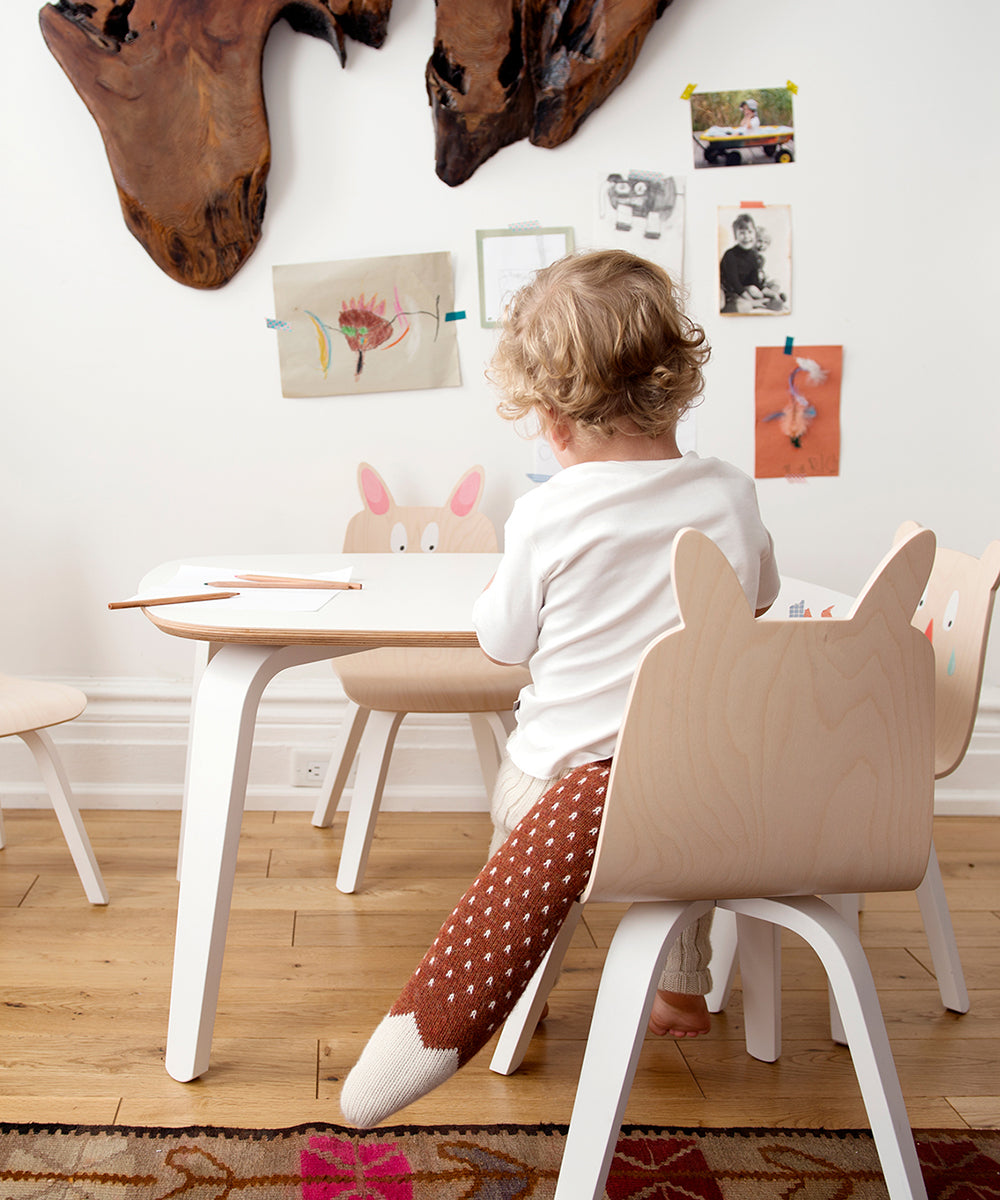 A child is drawing at a table while sitting on a Bunny Play Chair. Art decorates the wall.