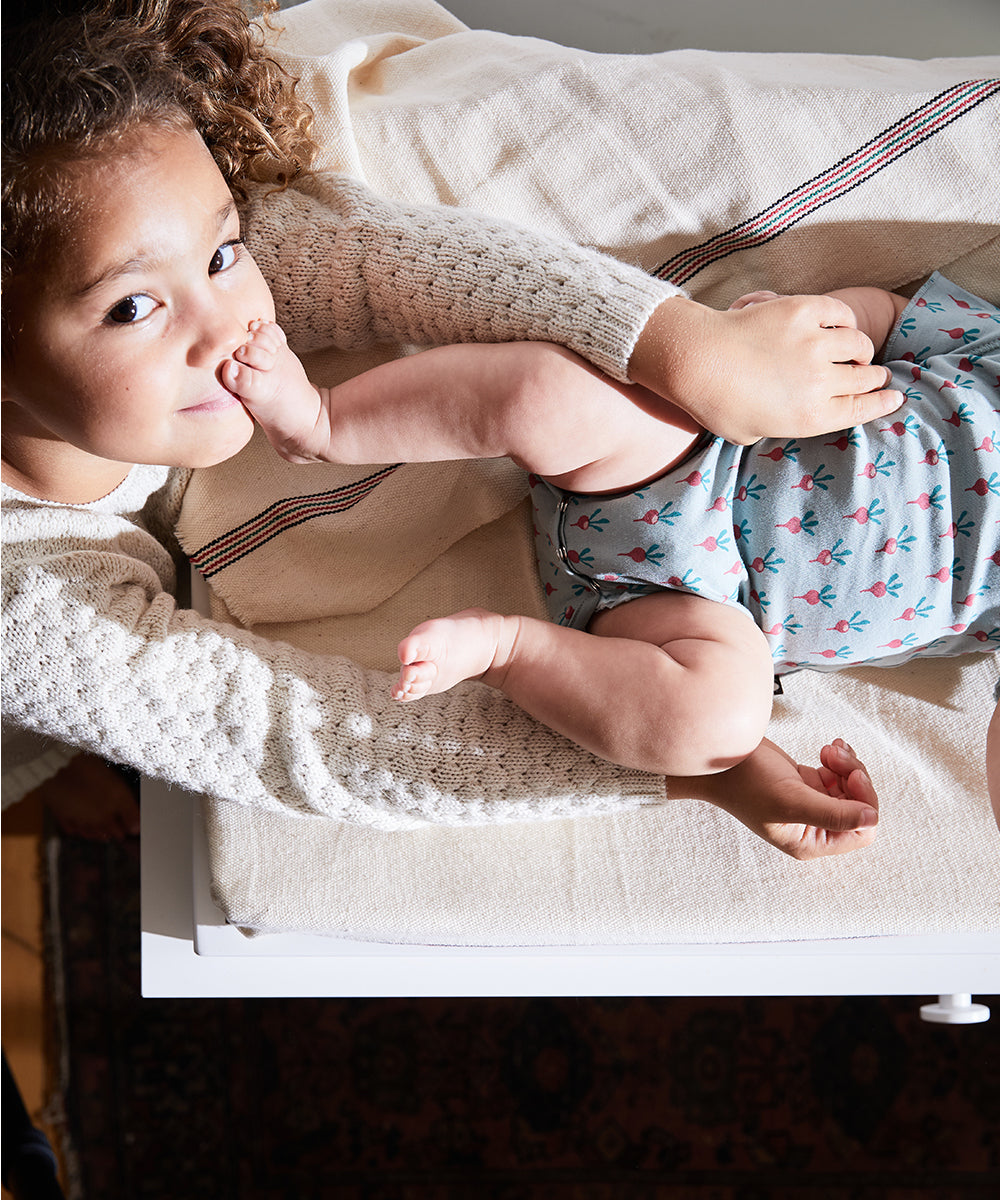 A young child playfully holds a baby in floral clothing, lying on a cream-colored Naturepedic 4-Sided Changing Pad.