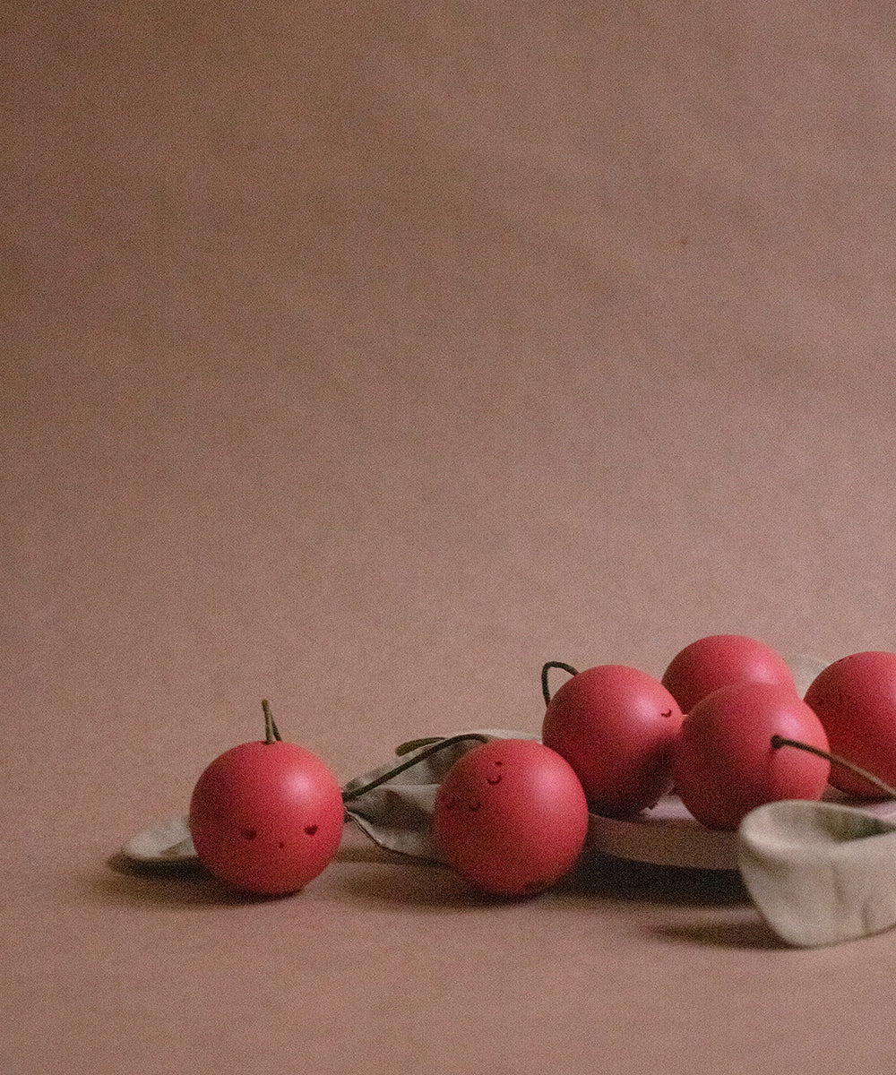 The Le Wooden Cherry Play Set features small red cherries with smiling faces on a beige surface, some placed on a plate, along with two nearby leaves.