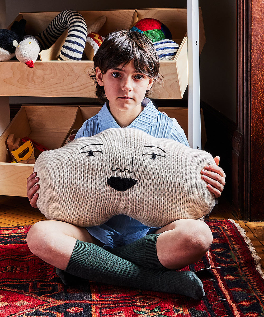 A child sits on a rug, holding a Cloud Pillow with a face, surrounded by toys on shelves.