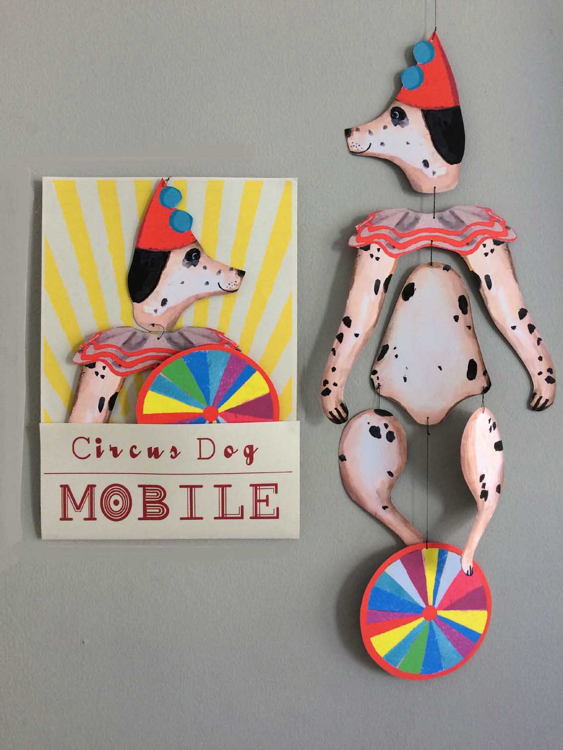 A Dalmatian in a red hat holds a colorful wheel in the circus-themed Le Mobile - Circus Dog, displayed next to its matching package.