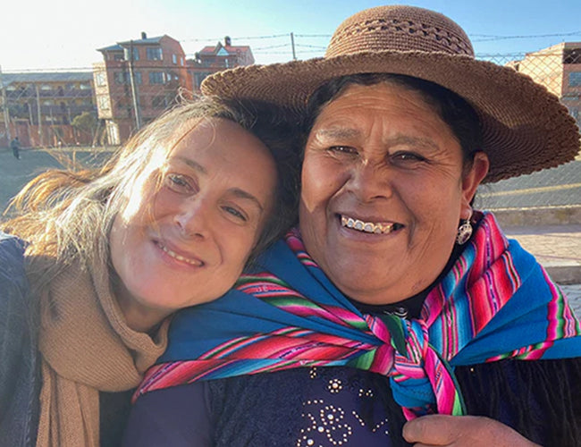 Two people smiling; one wearing a scarf, the other in a patterned shawl and hat. Theyre outdoors with buildings in the background.