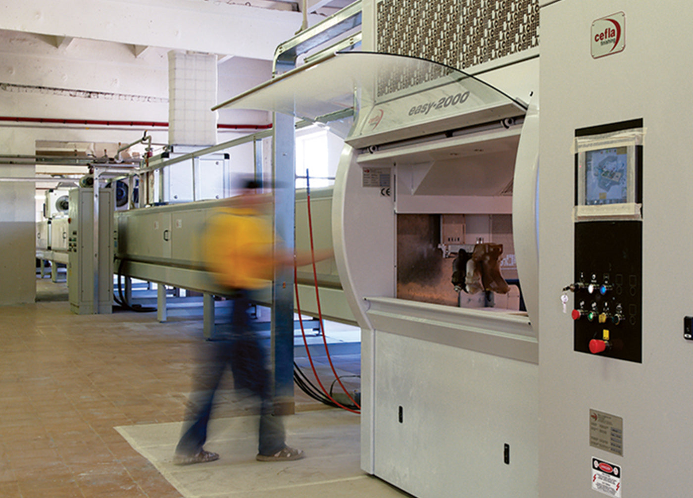 A person in motion stands near the Cella Easy 2000 machine in a large industrial space with equipment and tiled flooring.