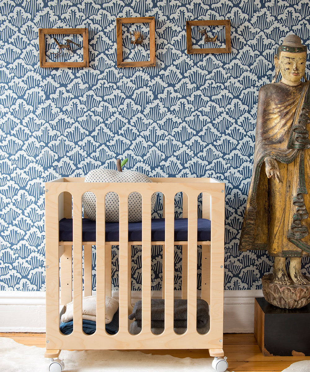 A Fawn 2-in-1 Crib System is in a room with blue patterned wallpaper, a small plant, framed pictures, and a statue.