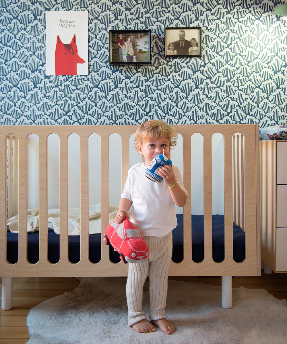 A toddler stands in front of a Fawn 2-in-1 Crib System, holding a red toy car and a drink, with patterned wallpaper and framed pictures in the background.