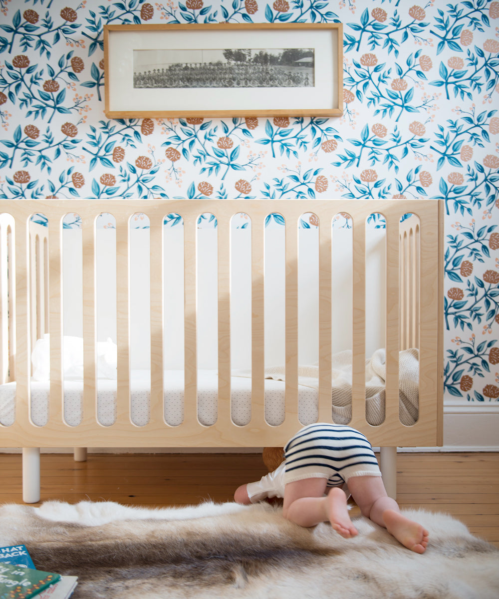 A baby crawls under the Fawn 2-in-1 Crib System in a nursery adorned with floral wallpaper and a framed picture on the wall.