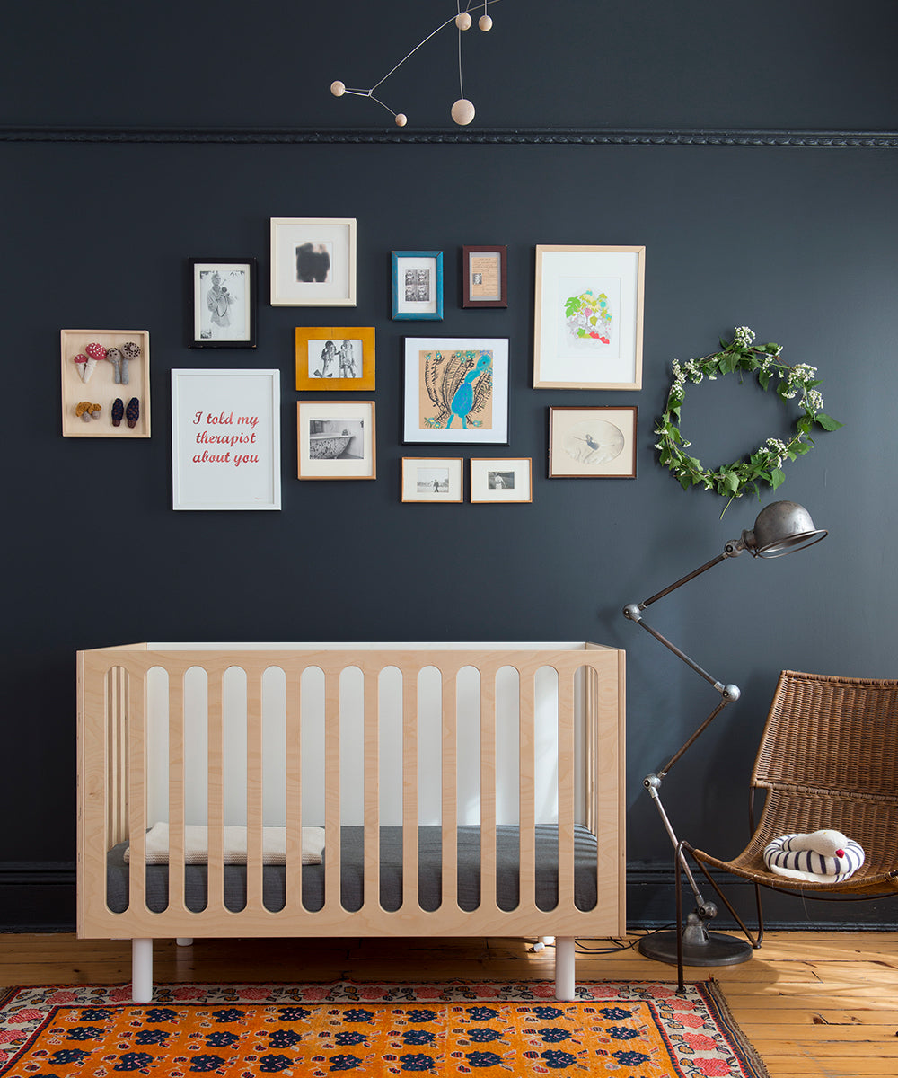 Contemporary nursery featuring the Fawn 2-in-1 Crib System in light wood, dark walls, a gallery wall, and a vibrant rug.