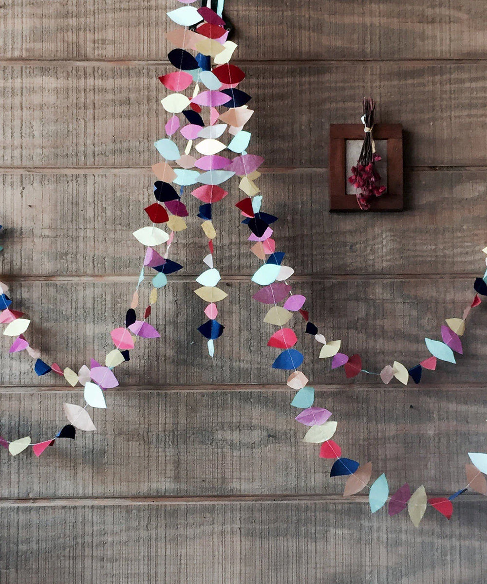 The Le Teardrop Garland adds a splash of color as it hangs on a wooden wall. A small framed picture with dried flowers complements the scene.