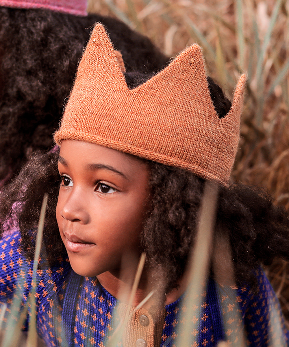 Child wearing a knitted gold crown and a blue sweater with orange patterns, sitting outdoors.