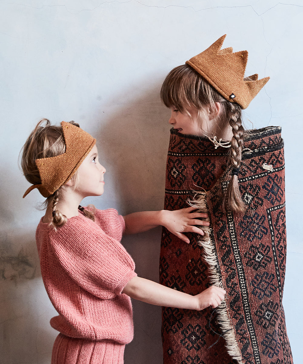 Two children playfully dressed; one wears a Gold Crown and is wrapped in a patterned rug, while the other dons a pink outfit.