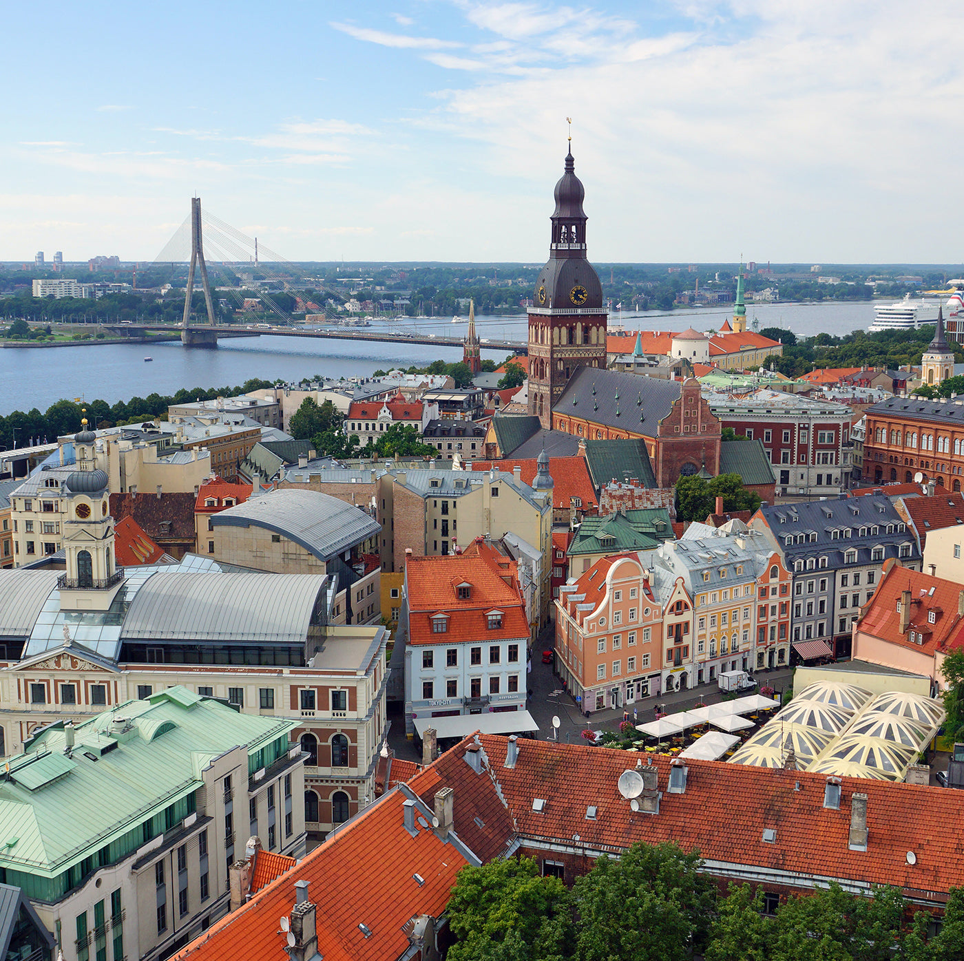 Aerial view of Riga, Latvia, featuring historic buildings, the Daugava River, a prominent church with a tall spire, and a bridge in the background under a partly cloudy sky.