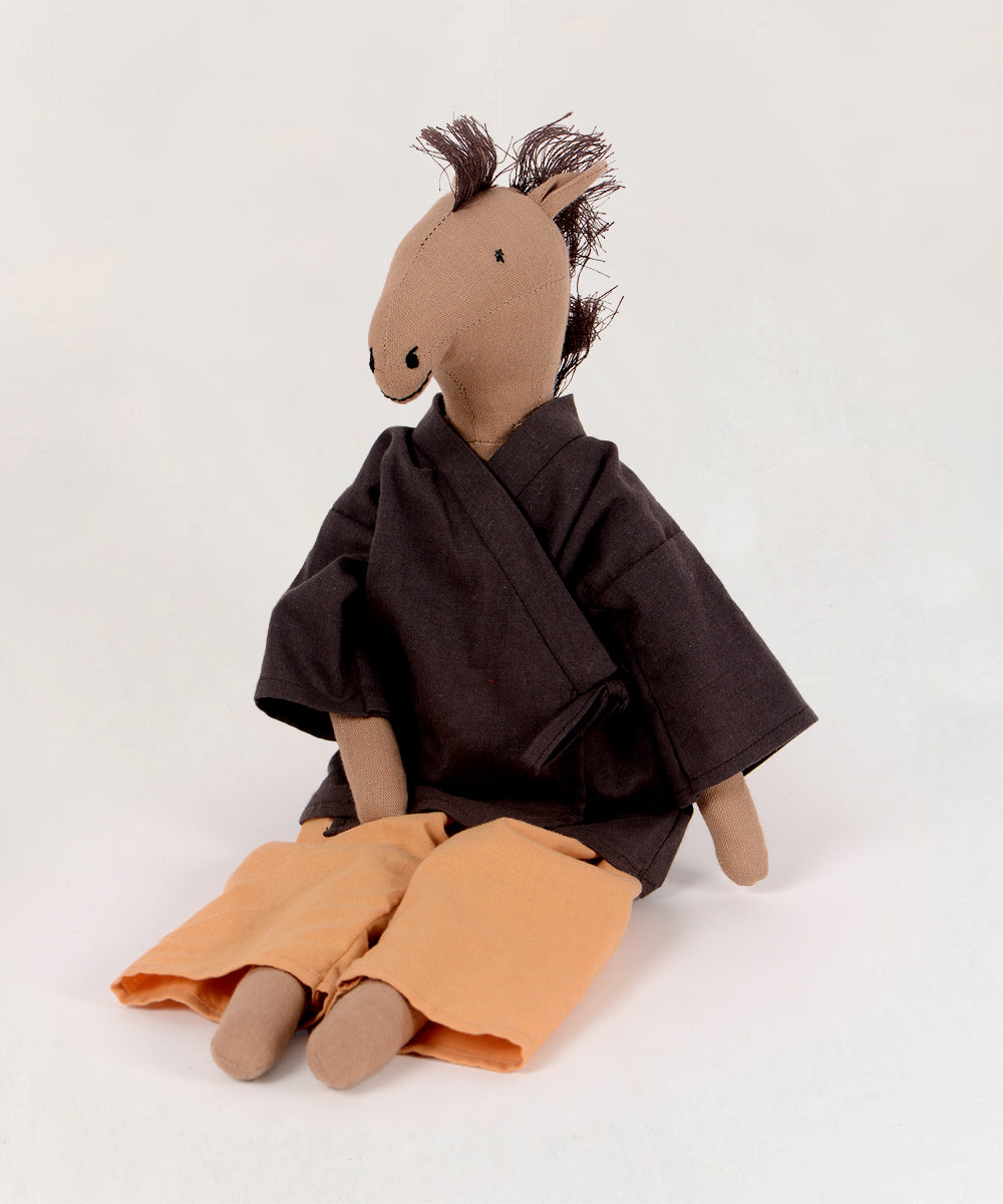 The Le Horsey doll, made of fabric, is dressed in a dark shirt and orange pants and sits against a plain background.