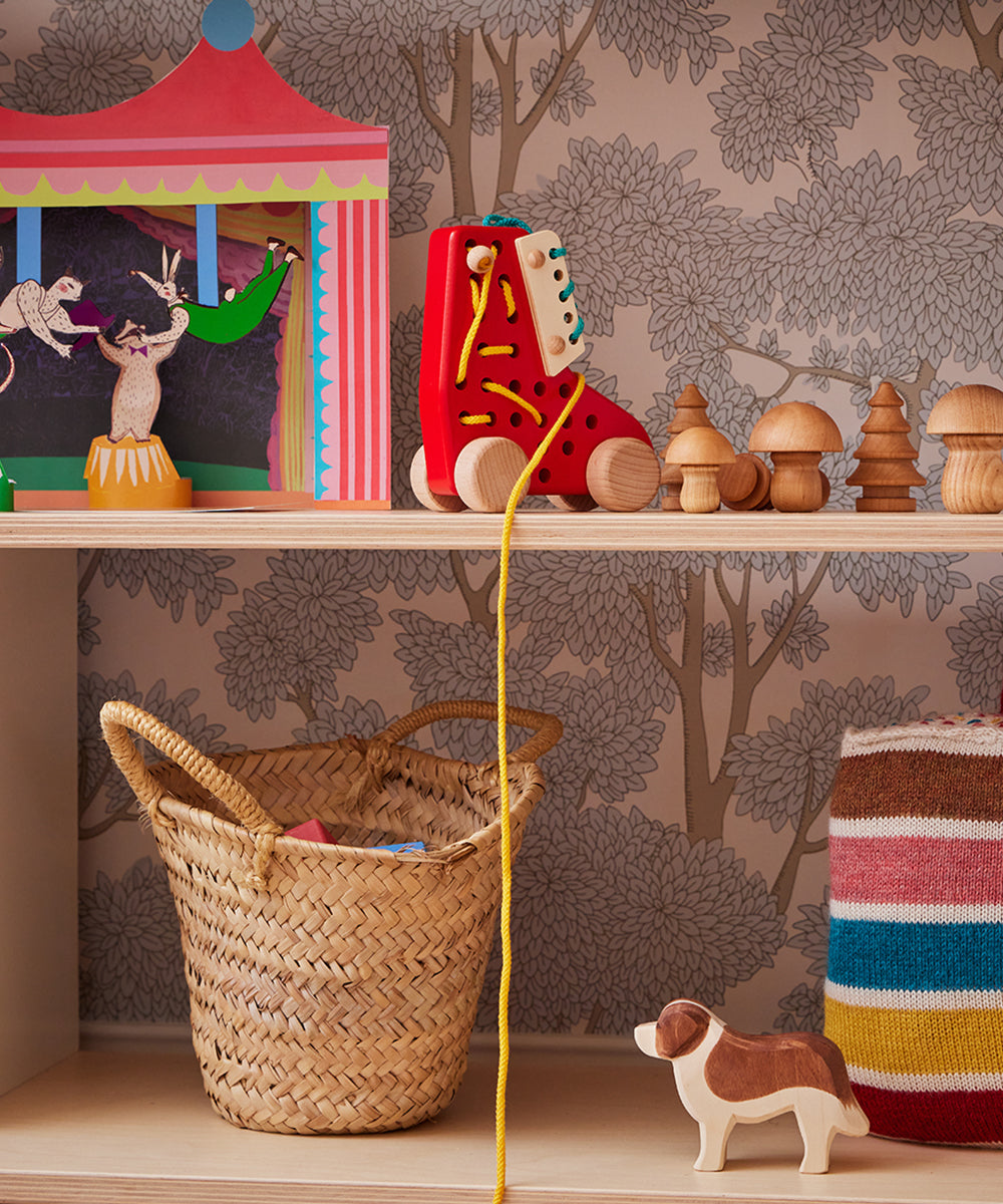 A shelf featuring a small Le Market Basket holding a toy theater, red lace-up boot toy, wooden mushrooms, wooden dog toy, colorful striped blanket, all against tree-patterned wallpaper.