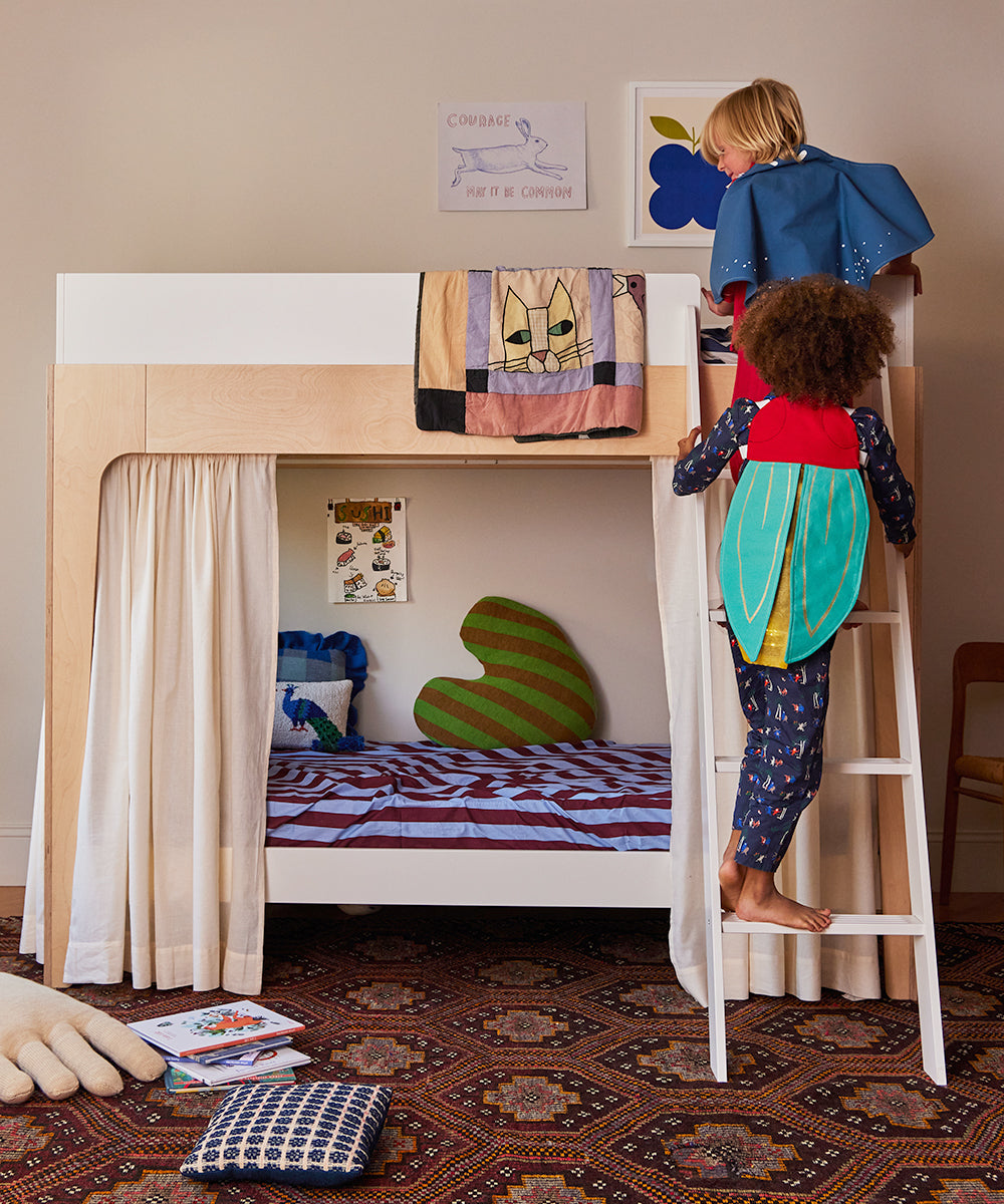 Two children wearing Le Firefly Costumes play on a wooden bunk bed with a curtain on the lower bunk, surrounded by posters, a rug, and toys.