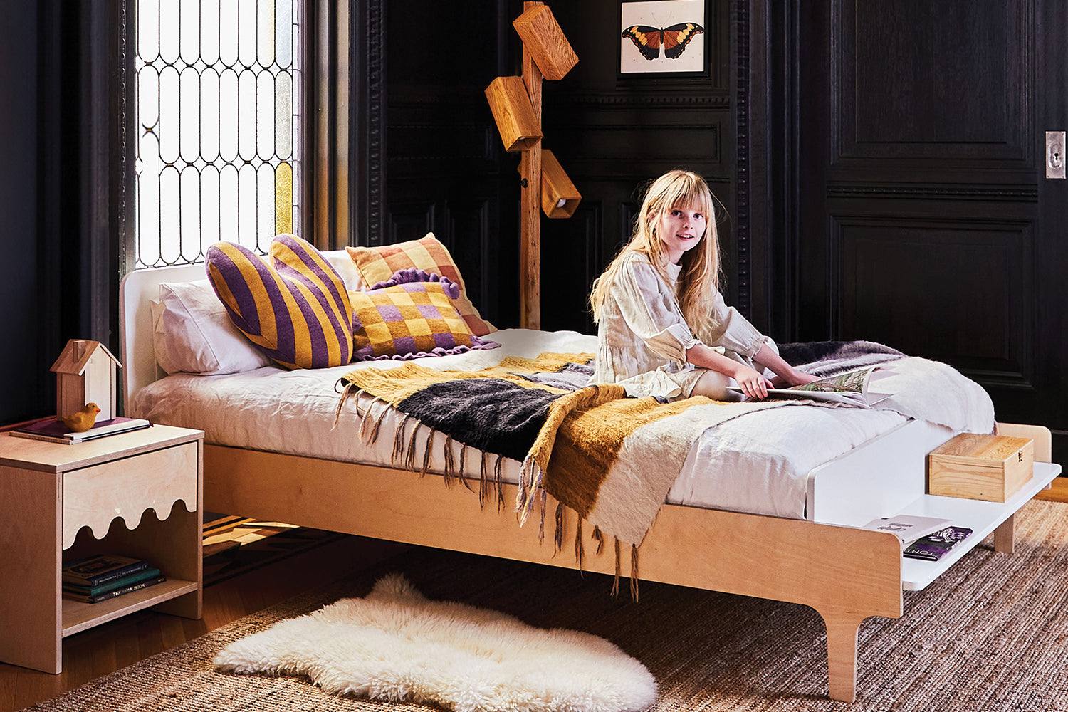 A woman sits on a modern wooden bed with layered blankets and cushions in a stylish bedroom. A plush rug, decorative items, and framed artwork complement the black walls and decorative window.