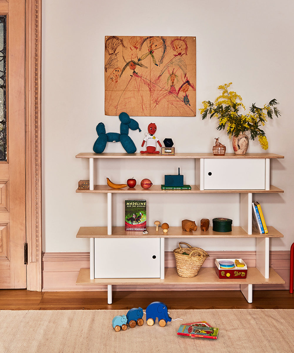 The Mini Library is adorned with toys, books, and decor. Above it hangs artwork, complemented by a vase of yellow flowers on the shelf.