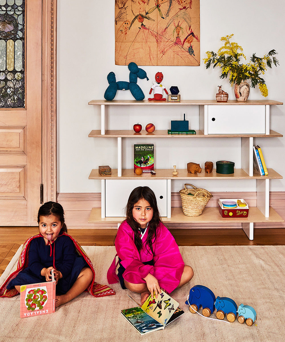 Two children sit on a rug with books. A Mini Library shelf behind them holds toys, plants, and books. Colorful artwork adorns the wall.