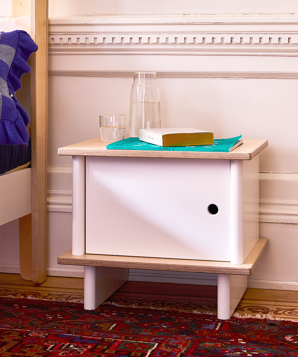 An ML Nightstand featuring a glass of water, carafe, and book on a turquoise mat is placed beside a bed with ornate wall molding in the background.