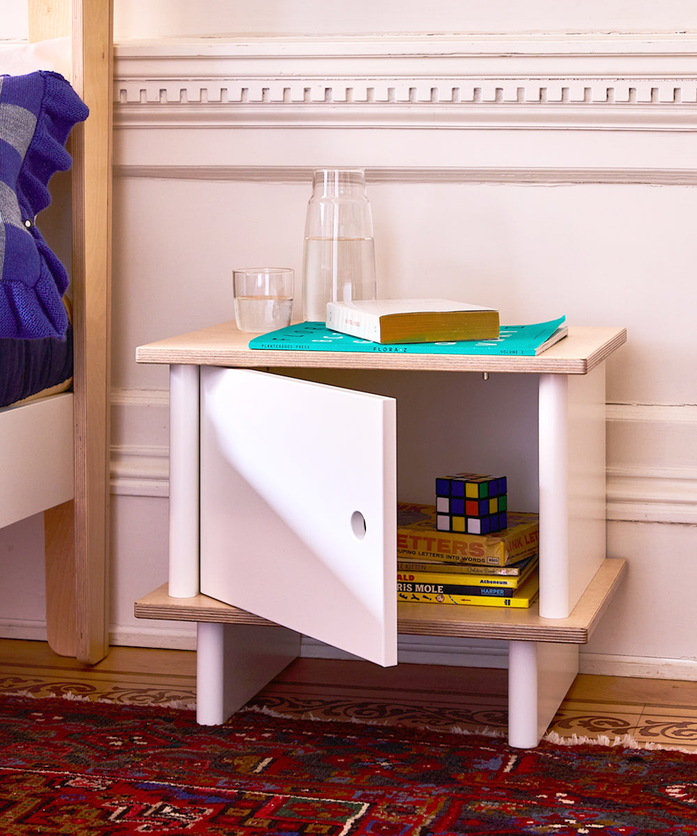 The ML Nightstand, with its door open, holds books and a Rubiks cube. On top are a glass, carafe, and another book.