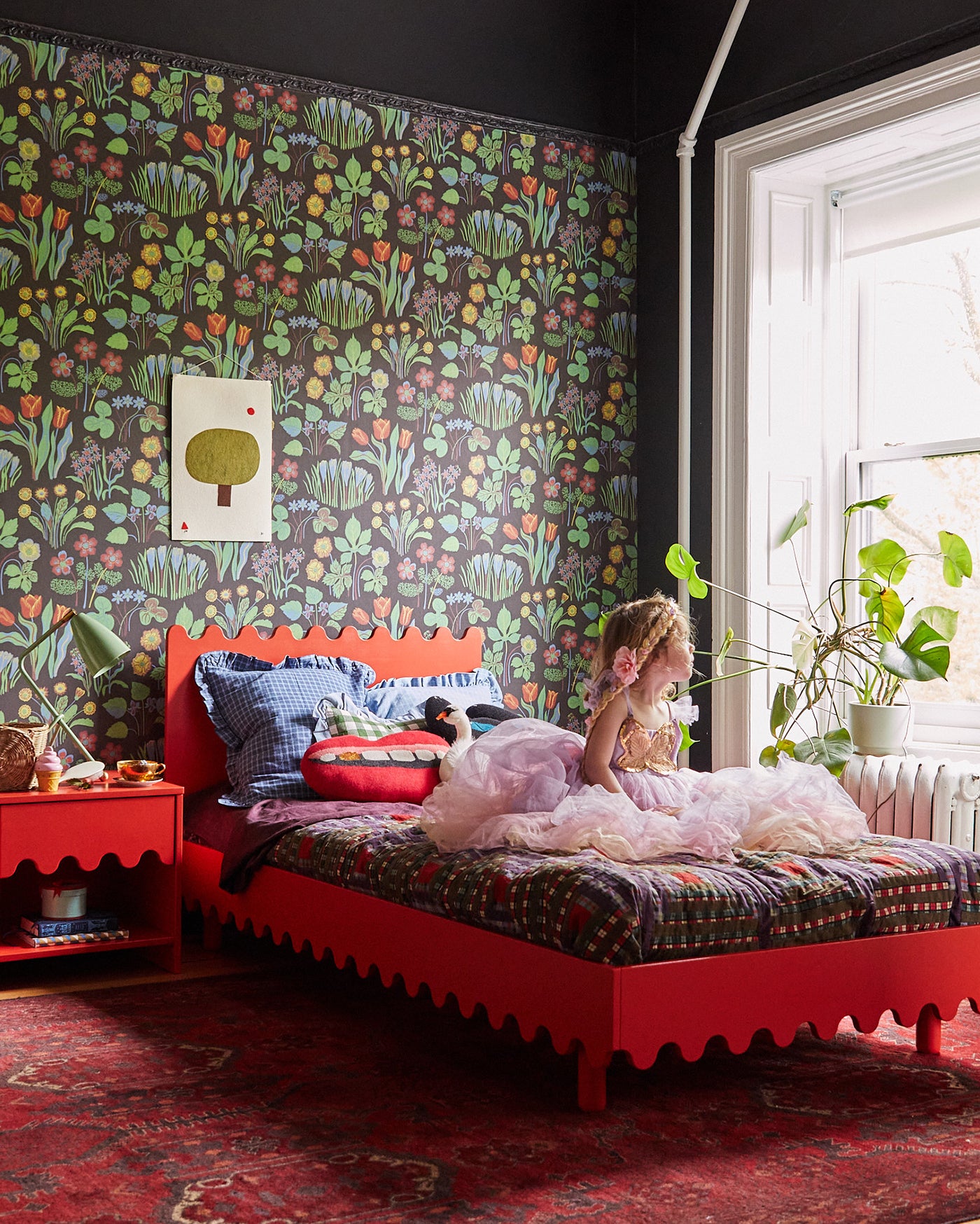 Child in a dress sits on a red bed in a colorful room with floral wallpaper, a plant by the window, and a red rug.