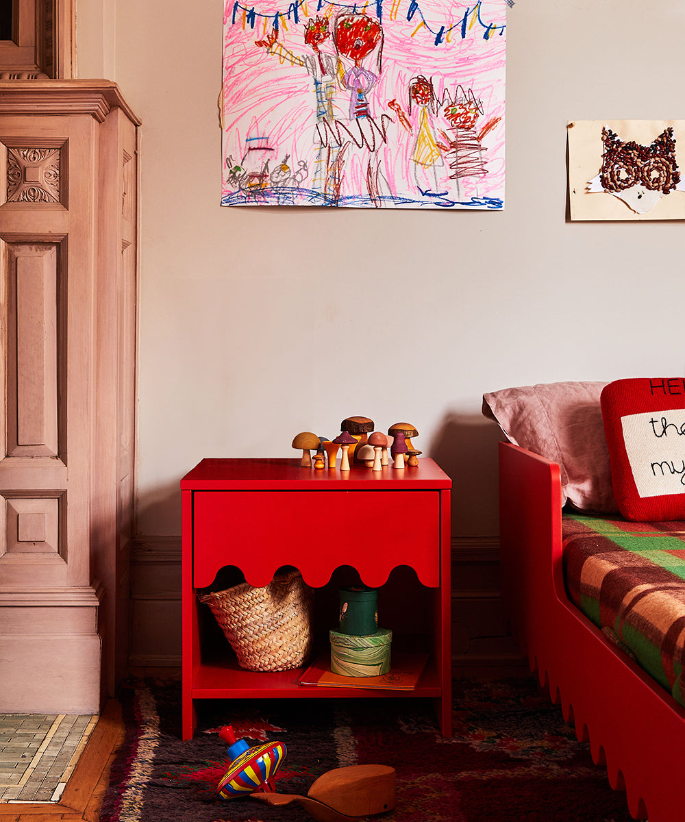 A cozy room featuring childrens art on the wall, a vibrant mix of toys atop a red Moss Nightstand, and a bed adorned with a colorful blanket.