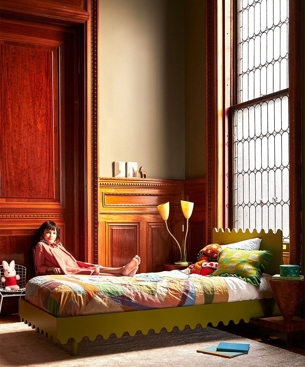 A child sits on a Moss Twin Bed with a multicolored quilt, beside a large window in a room with wooden paneling.