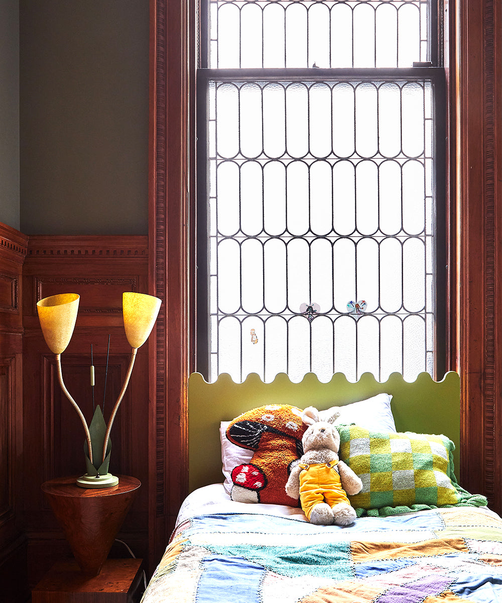 A childs vintage-style room features a Moss Twin Bed, colorful pillows, and a decorative lamp under a large window with patterned glass.