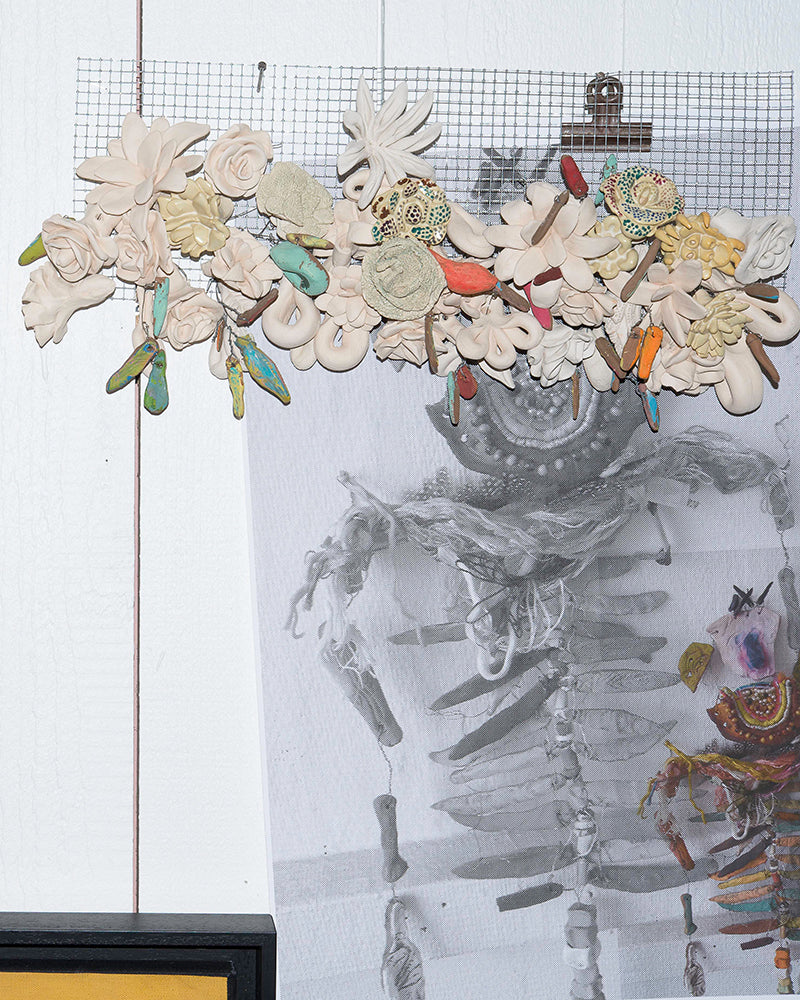 Art installation featuring a wire grid with assorted ceramic flowers and colorful objects above a grayscale skeletal-themed artwork on paper.