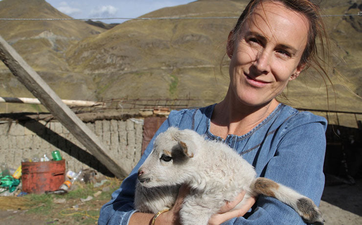 A woman in a denim shirt holds a small white and brown lamb outdoors in a mountainous area.