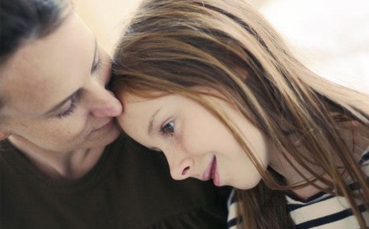 A woman gently rests her head against a young girl who is smiling. Both appear content and close together.