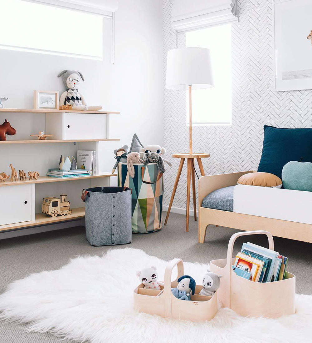 A cozy childs room with a rug, toy baskets, a shelf with books and toys, and a lamp. Neutral color scheme with white walls and natural wood accents.