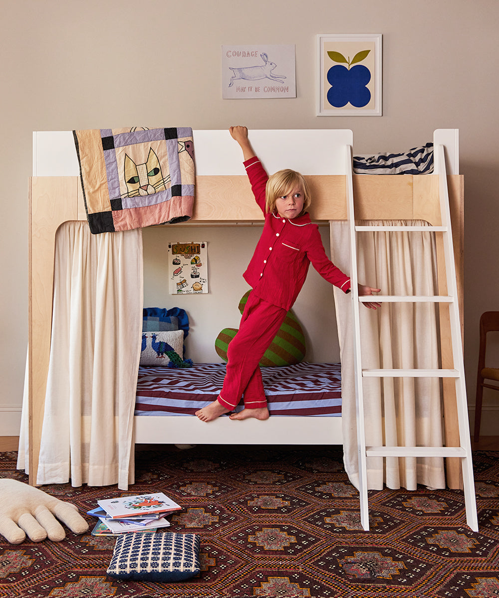 A child in red pajamas stands on the ladder of a Perch Twin Bunk Bed in a cozy room with books and toys scattered on a patterned rug.