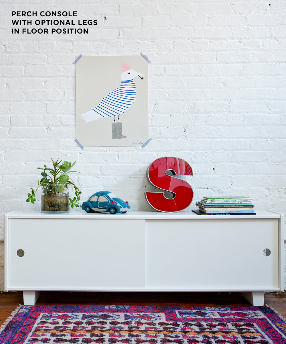 The Perch Console - Full Size features books, a red S, toy car, plant, and seagull art on a brick wall above a patterned rug.