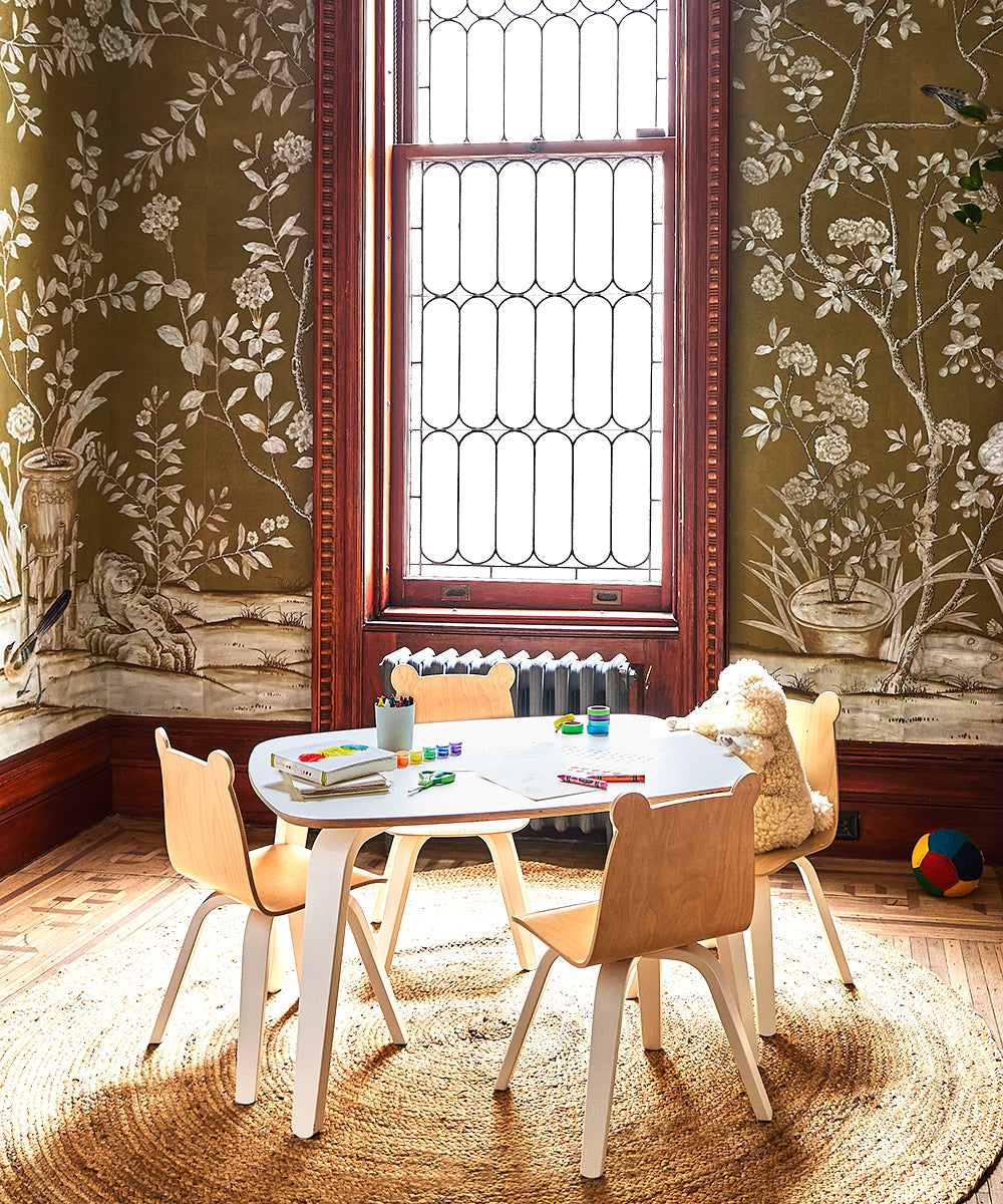 The kid-sized space includes a playroom with a Play Table, four chairs on a round rug, and ornate wallpaper. A teddy bear occupies one chair, art supplies are spread across the table, and a colorful ball rests on the floor.