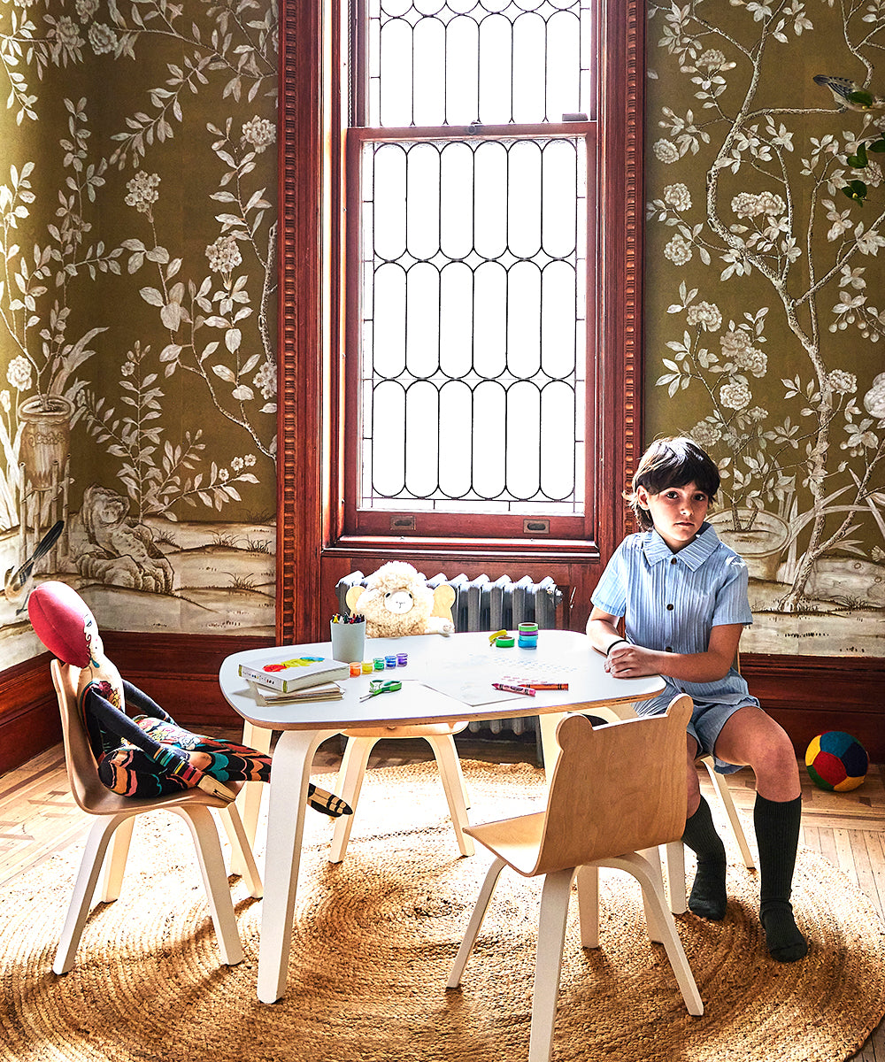 A child sits at a Play Table in a cozy, kid-sized area, surrounded by toys and a colorful ball. The rooms intricate floral designs and large stained glass window are all crafted from sustainable materials.