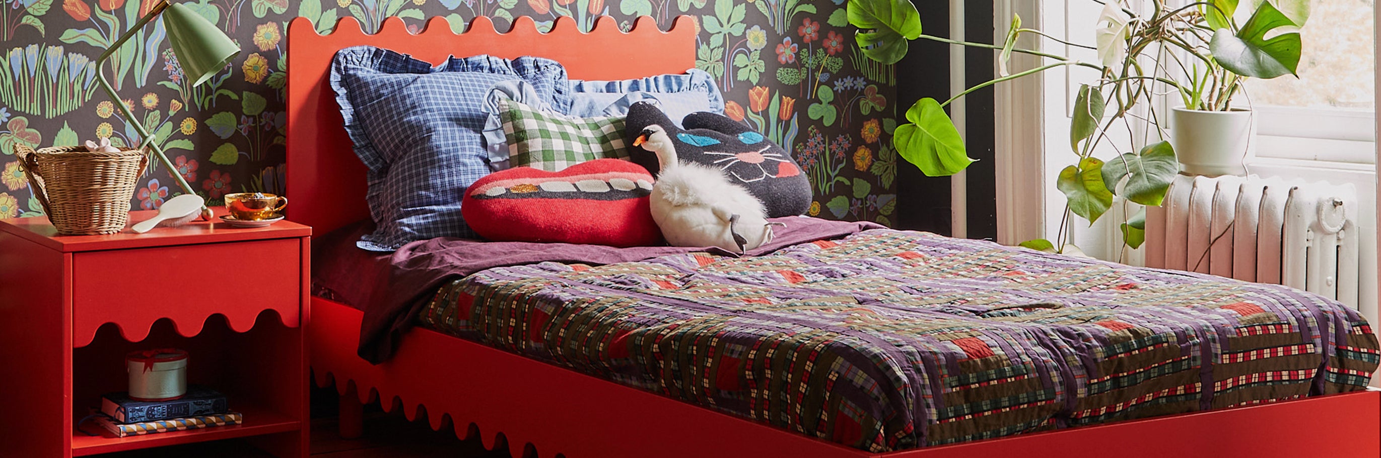 A bedroom with a red bed, plaid bedding, and various pillows. A red nightstand holds a plant and basket. The room has patterned wallpaper and a large leafy plant by the window.