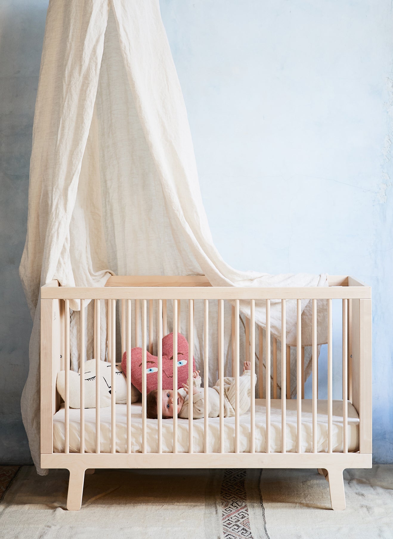 A wooden crib with white bedding and a hanging canopy. Inside are stuffed toys: a pink rabbit and a white bear. The background wall is light blue.