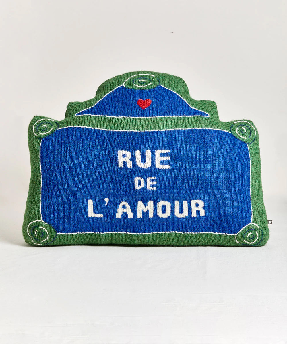 The Rue de lAmour Pillow is a knitted pillow that resembles a street sign, featuring Rue de lAmour in white on a blue and green background.