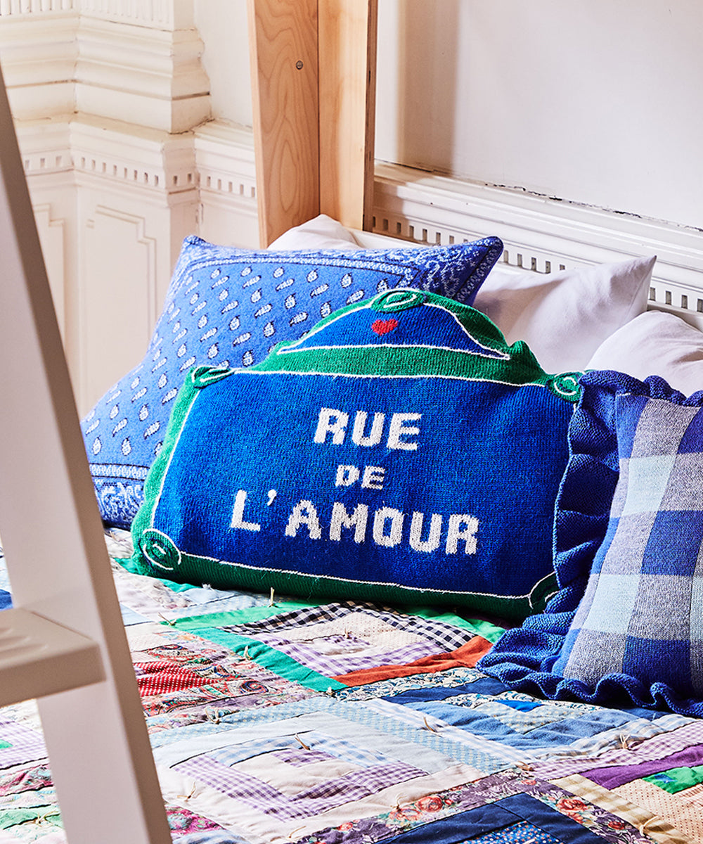 Three decorative pillows on a bed, featuring one labeled Rue de lAmour in blue and green, are set against a quilted blanket.