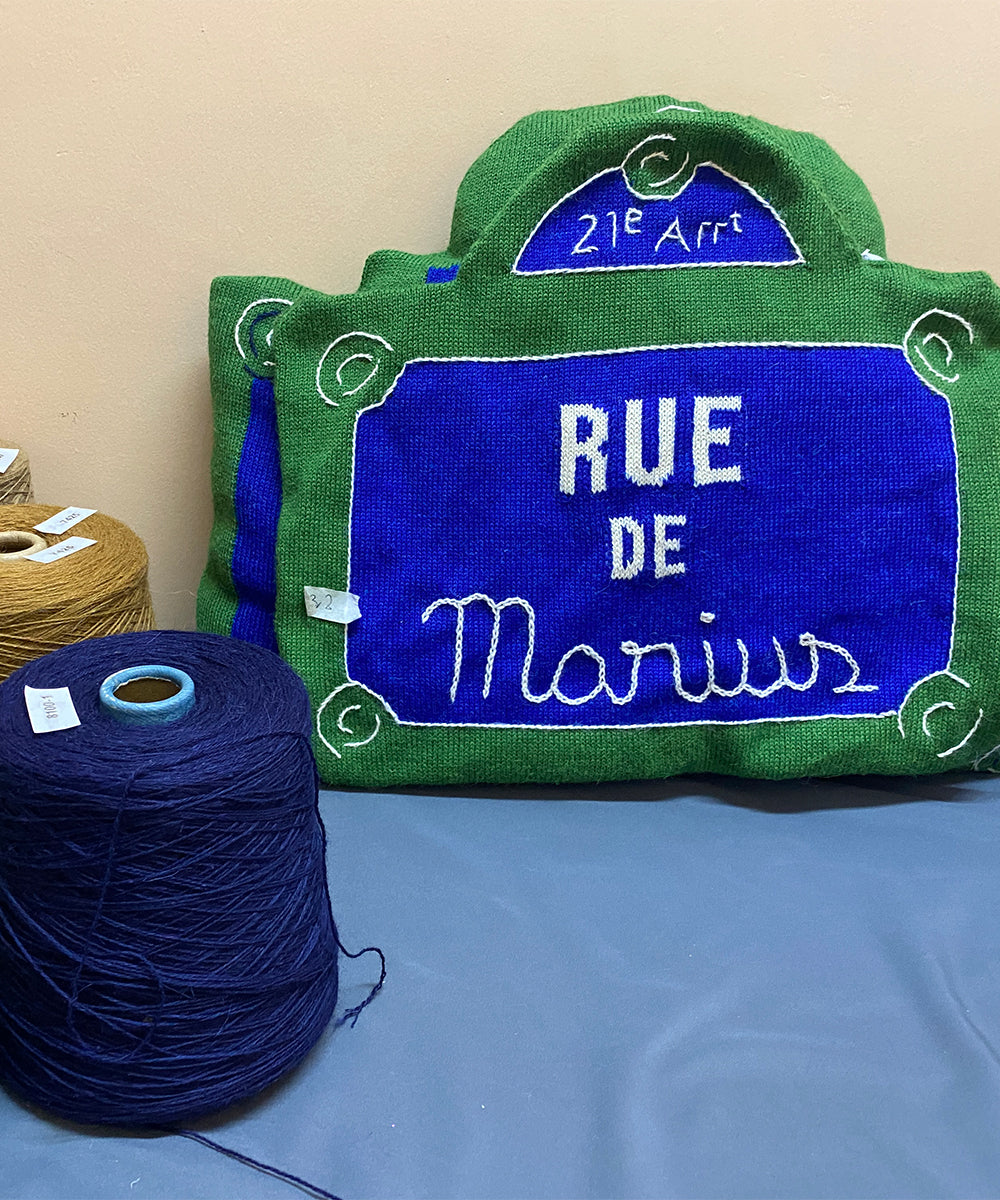 Knitted pillow called Rue de lAmour next to spools of brown and navy yarn, resembling a street sign.