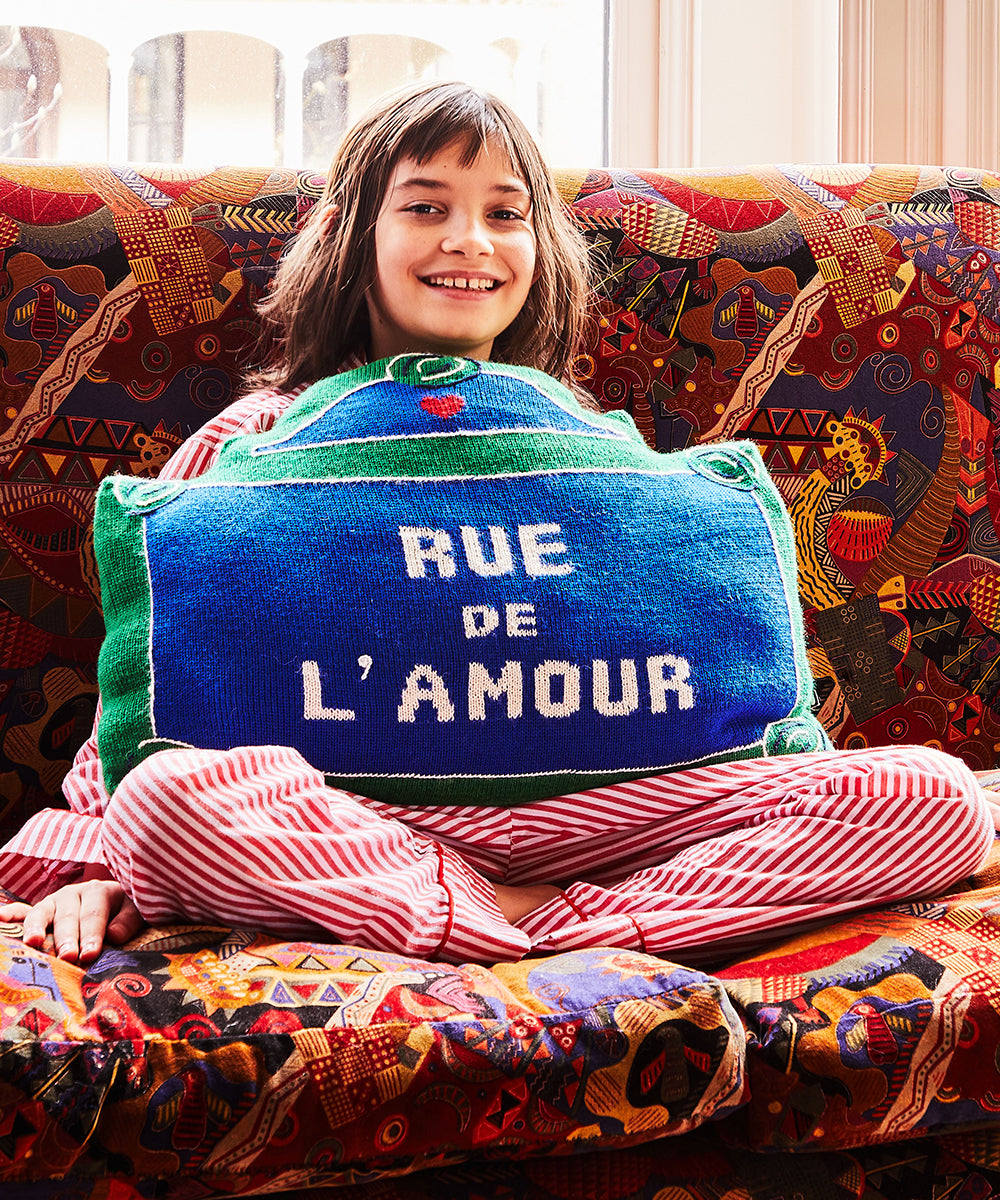 A child in striped pajamas sits on a colorful sofa, holding a Rue de lAmour Pillow.