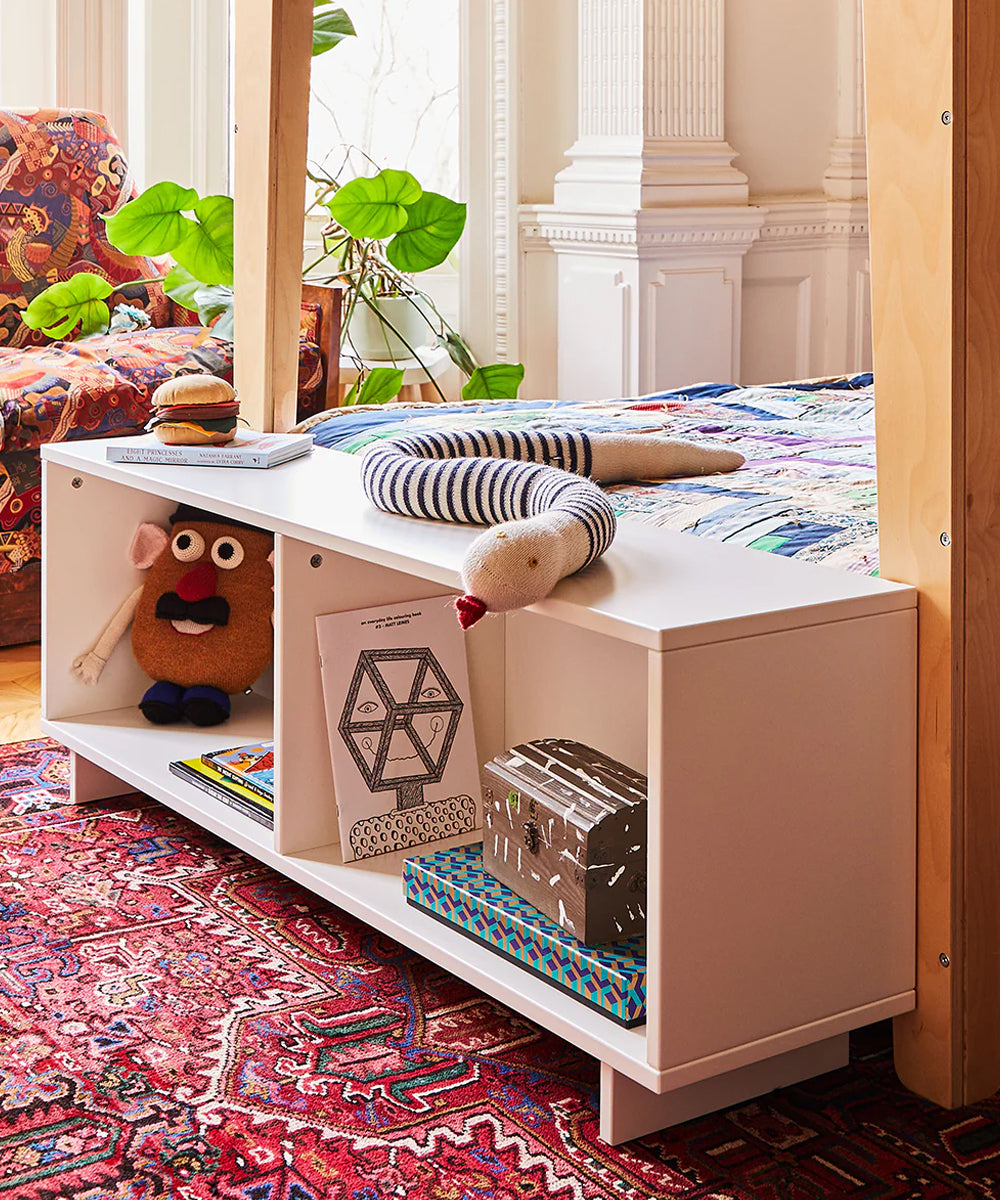 Snake Pillow at the foot of a bed, with toys and books on a white shelf; nearby, there's a colorful patterned rug and a plant.