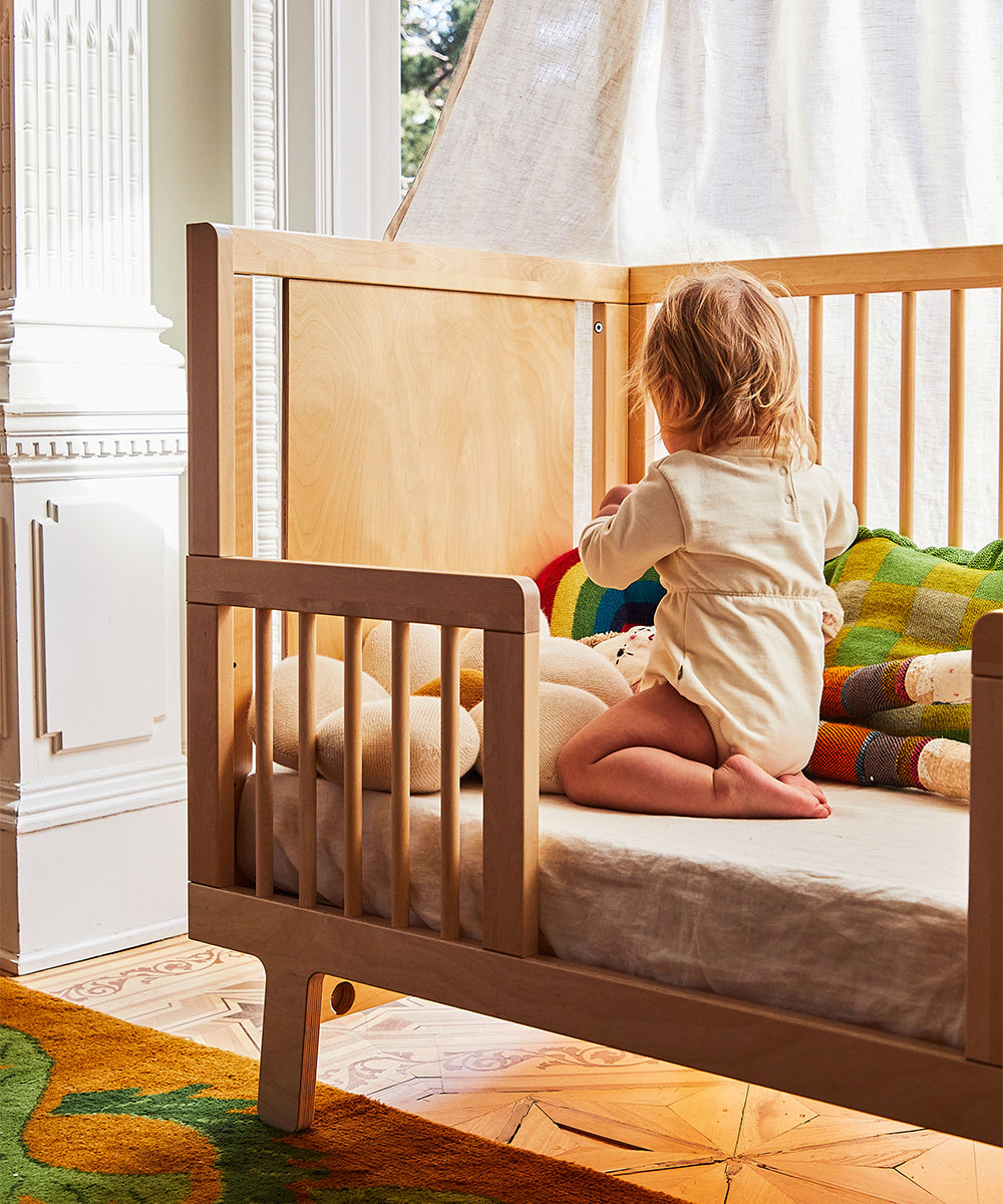 A child kneels and plays with toys and blankets in a crib enhanced by the Sparrow Toddler Bed Conversion Kit, as daylight streams through a nearby window.