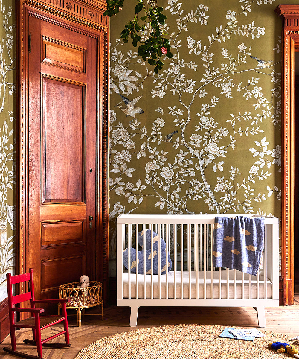 Cloud Blanket draped over a white crib in a floral wallpapered room, with a wooden door, red chair, and plants above.