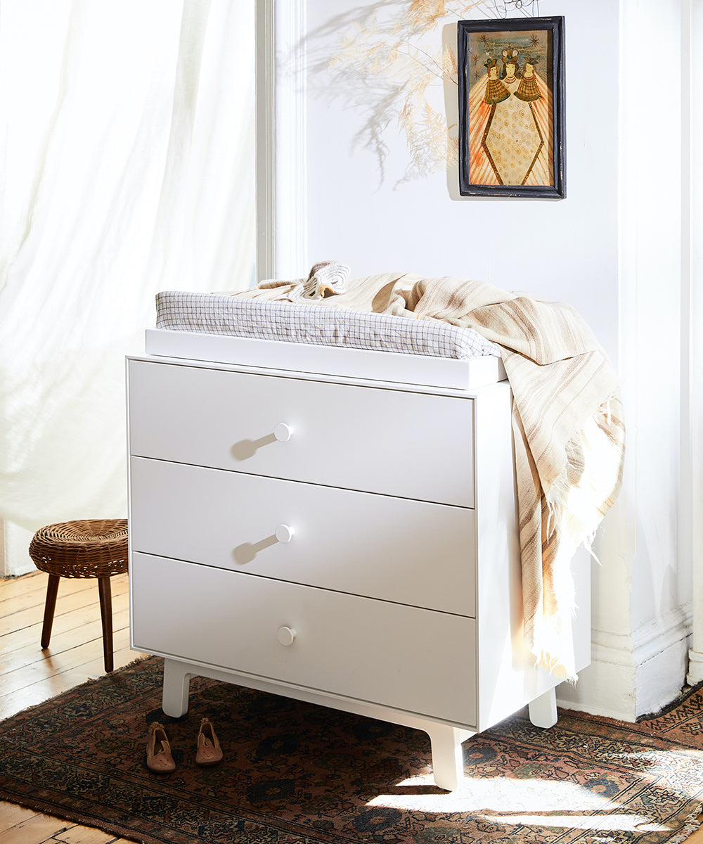 White dresser with three drawers, a Naturepedic 4-Sided Changing Pad, and striped blanket; art hangs above; shoes and rug on the floor.