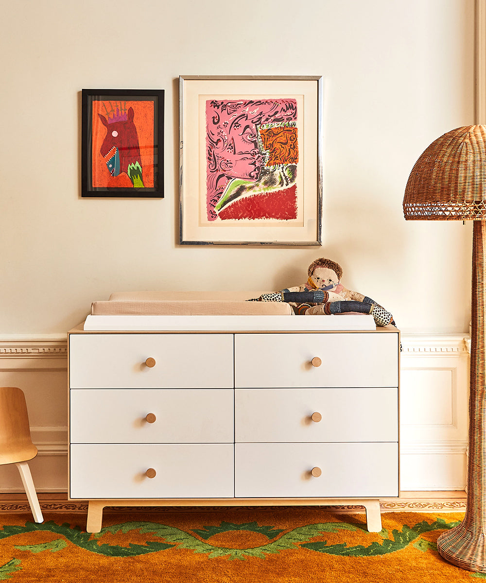 The Convertible XL Changing Station, adorned with wooden knob handles, sits beneath two framed artworks, holding nursery essentials like a plush doll. A wicker lamp and wooden chair are on either side, while an orange patterned carpet adds warmth to the ethically crafted space.