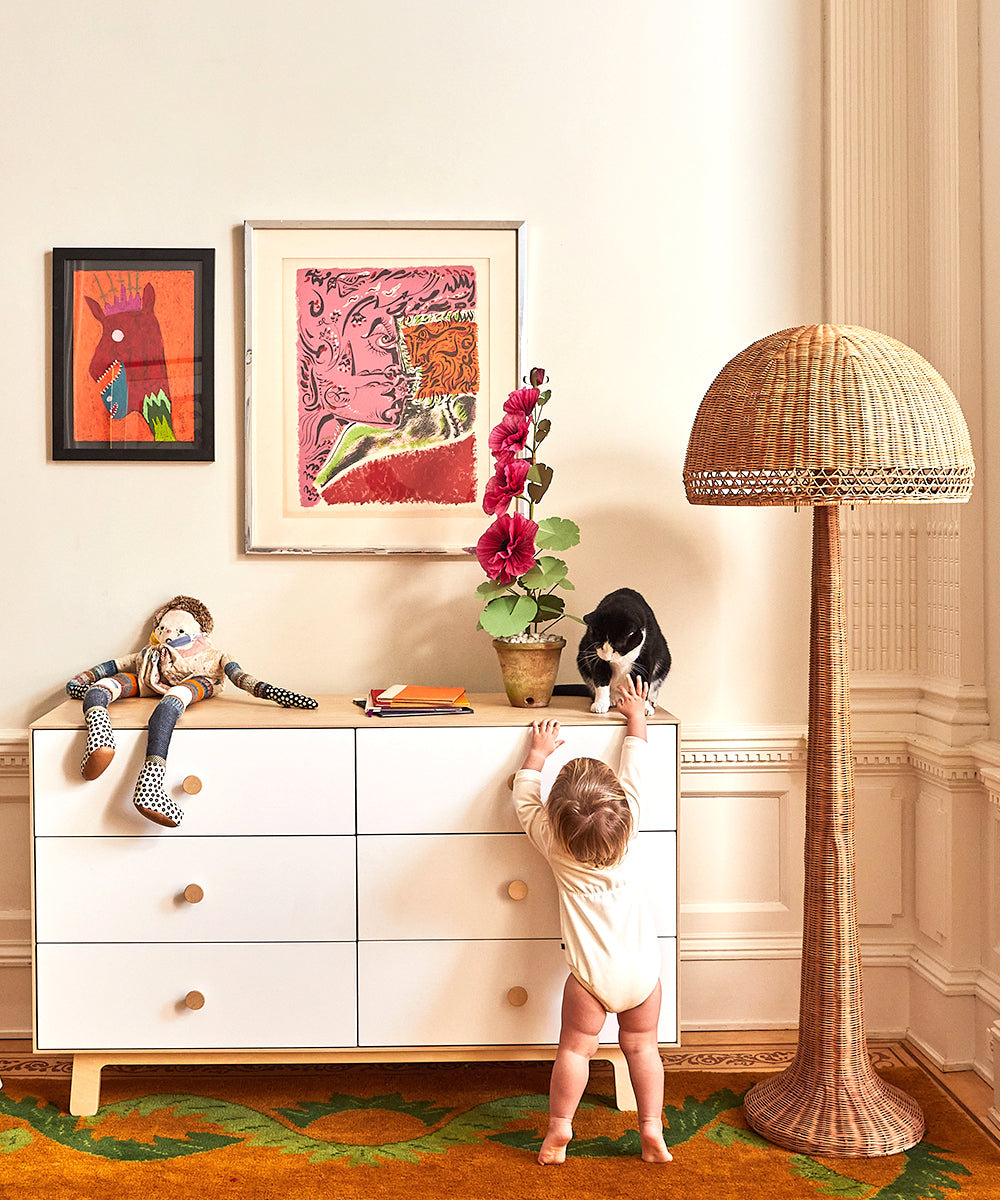 A baby reaches up to the Sparrow 6-Drawer Dresser where a cat sits atop; art and a lamp adorn the background.