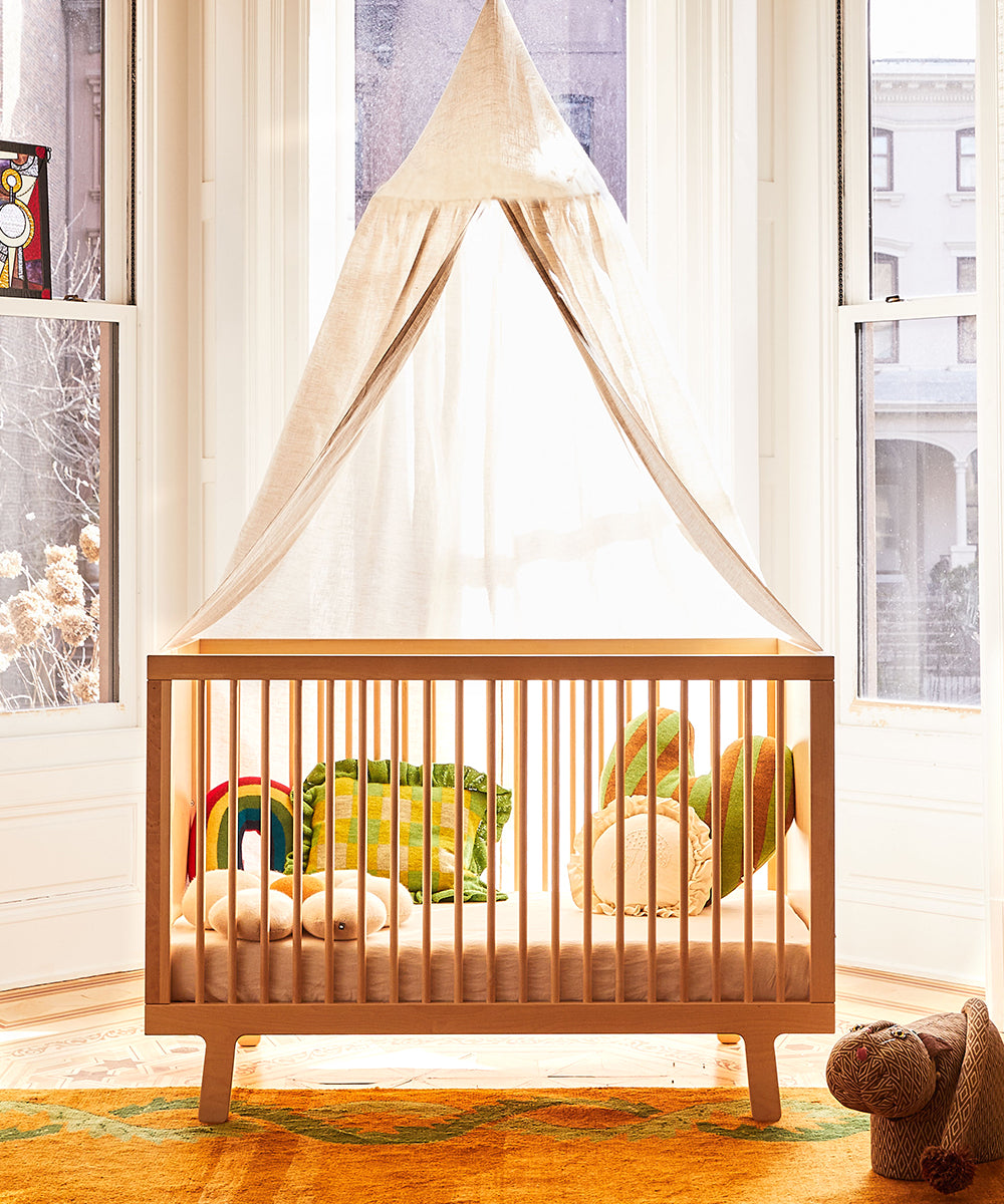 The Sparrow Crib, crafted from birch hardwood and GREENGUARD Gold certified, is situated in a bright room with plush toys, pillows, and a rainbow decoration. Large bay windows illuminate the space.