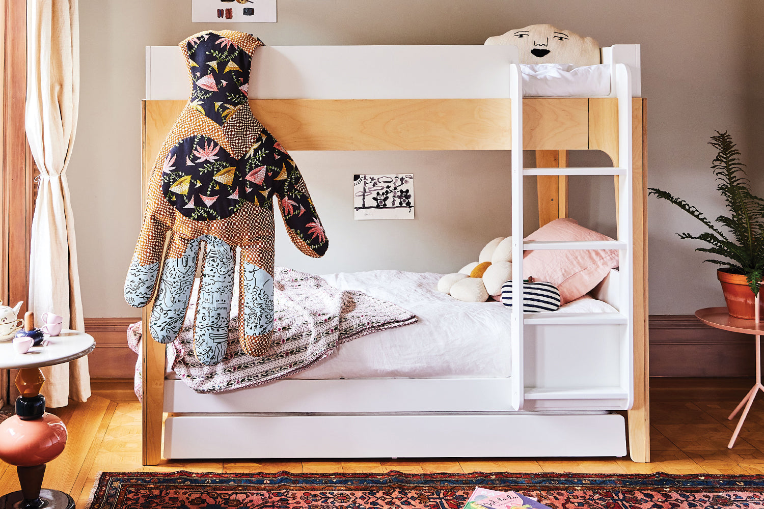 A childrens room with a wooden bunk bed, colorful quilt, plush toys, and a small side table. A plant is placed in the corner, and light streams in through large curtains.