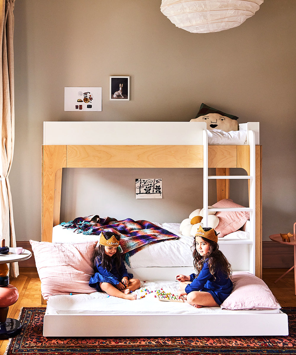 Two children in crowns sit on a Perch Trundle Bed, part of the Ladder Conversion Kit, playing with toys in a cozy bedroom adorned with vibrant bedding.